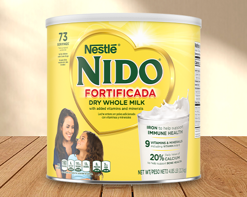 Nestlé Nido Fortificada dry whole milk container on a wooden surface against a warm beige background. / Envase de leche entera en polvo Nestlé Nido Fortificada sobre una superficie de madera y un fondo beige cálido.