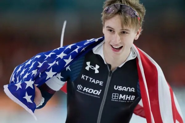 Jordan Stolz, a 21 year old American speed skater, grins while holding an American flag over his shoulders.