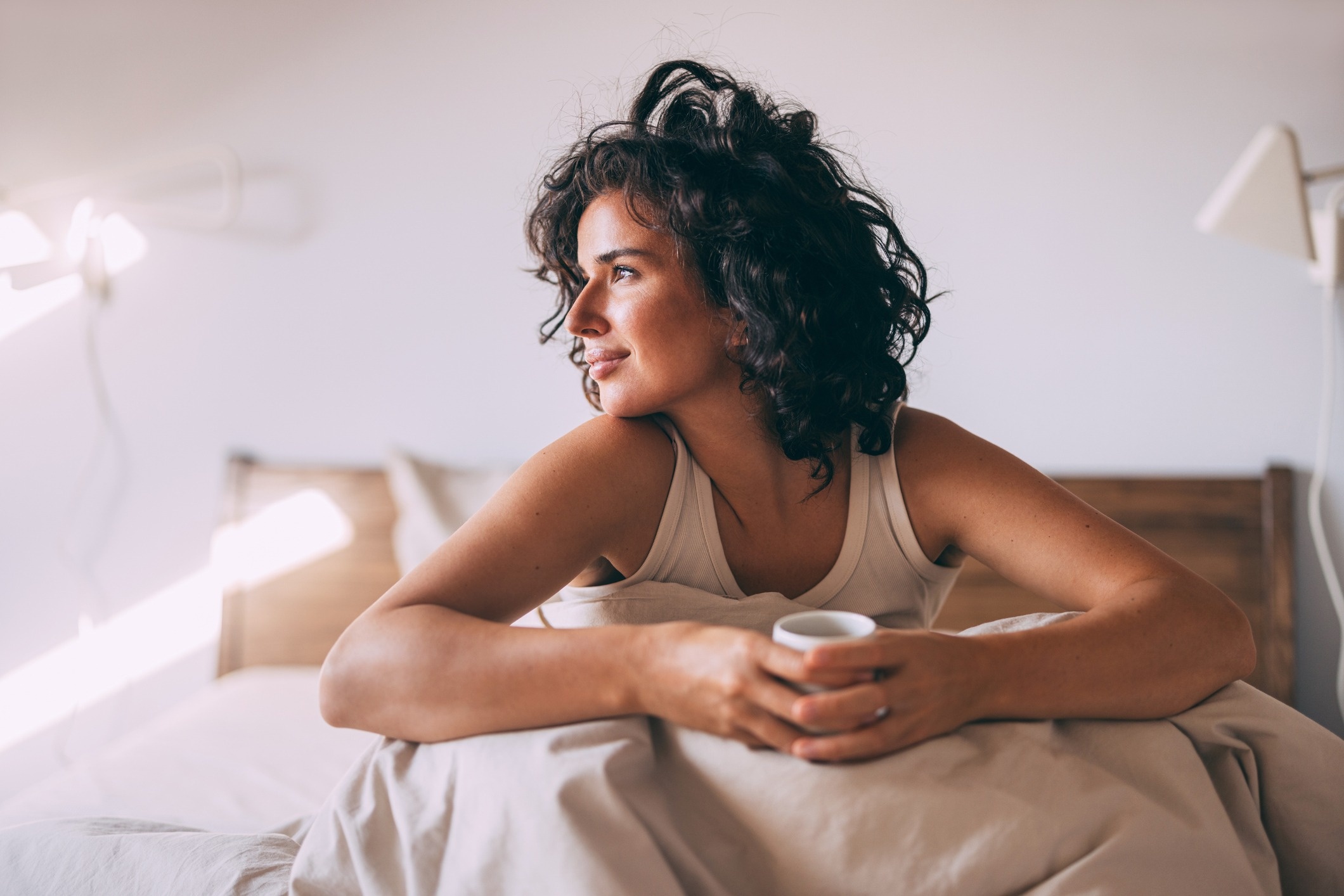 Woman sitting up in bed, holding a mug and looking out toward sunlight, with a relaxed expression in a softly lit bedroom.