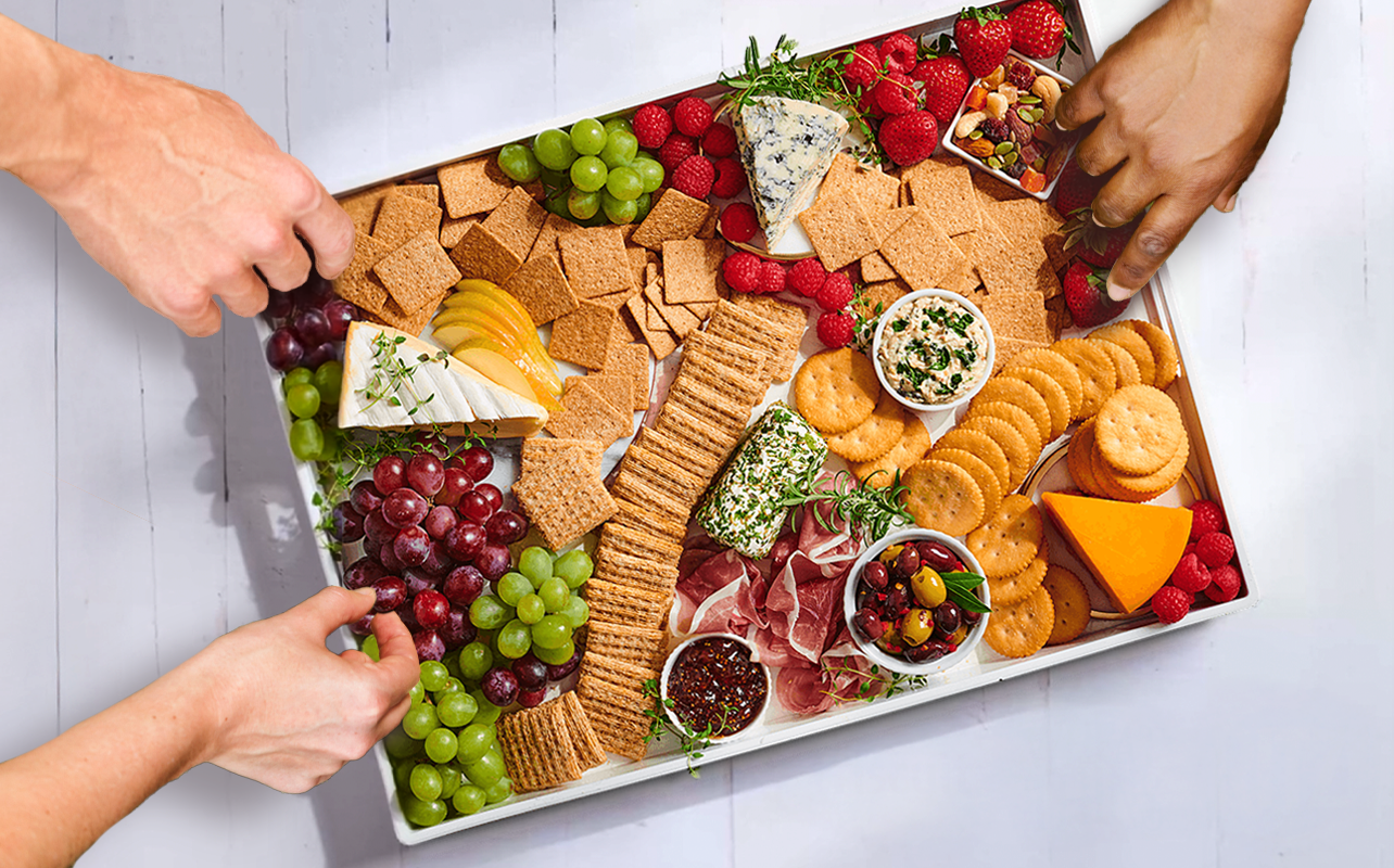 Board of crackers, cheese and fruit with hands reaching for snacks.