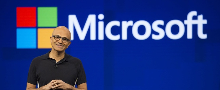 Satya Nadella presenting on a stage in front of a large blue backdrop featuring the Microsoft logo.