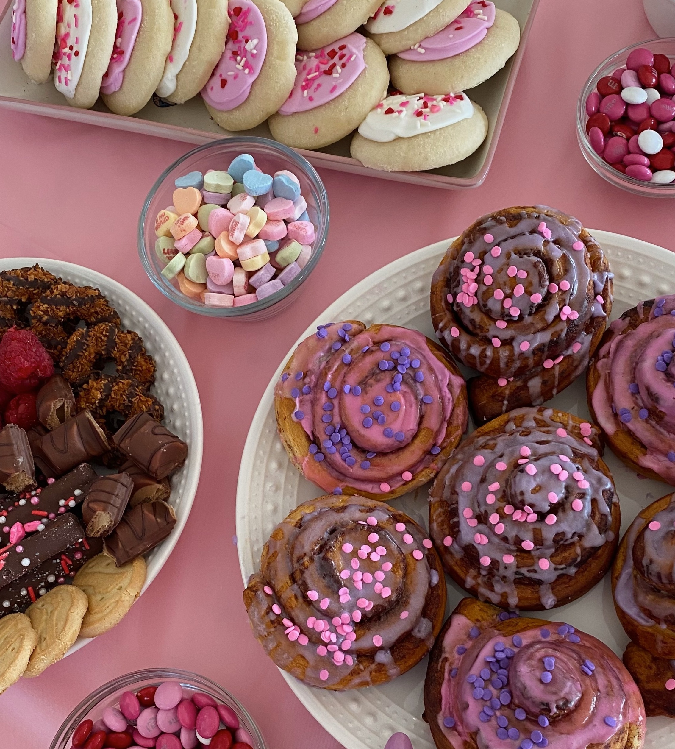 Assorted pink and white frosted cookies, cinnamon rolls with sprinkles, and bowls of candy arranged on a pink tablecloth.