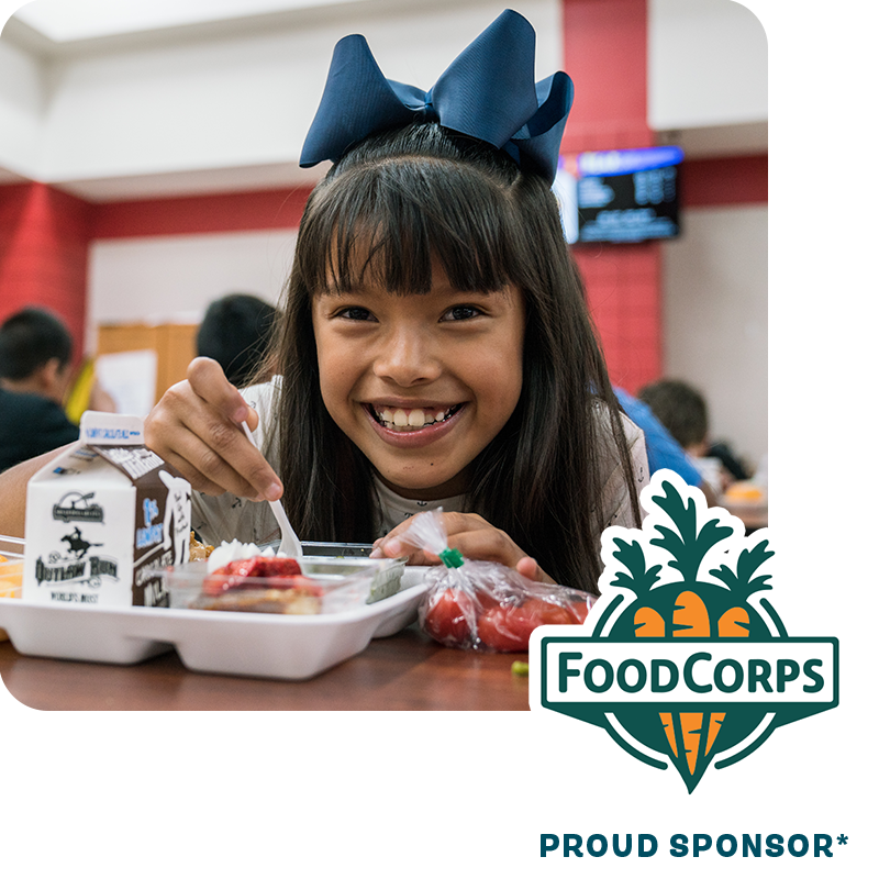 Girl sitting at a table in a cafeteria eating lunch with the FoodCorps logo;