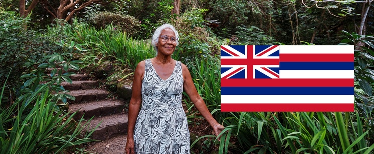 An elderly woman standing on outdoor stone steps surrounded by lush tropical greenery, with the flag of Hawaii overlaid on the right.