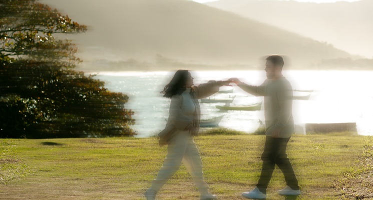 A couple dances together on a grassy shore at sunset, with water, boats and hills in the background.