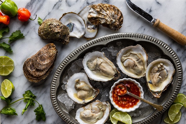 A view of fresh oysters on ice in a silver tray, surrounded by whole oyster shells, lime wedges, peppers, and a small dish of red mignonette sauce on a marble countertop.
