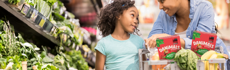Girl and parent in a grocery store fresh vegetable aisle with Danimals products in shopping cart.