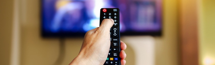 A hand holds a TV remote up in front of a blurred TV screen.
