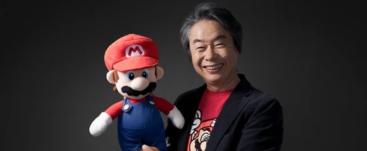 Shigeru Miyamoto smiling alongside a large plush toy of Mario.