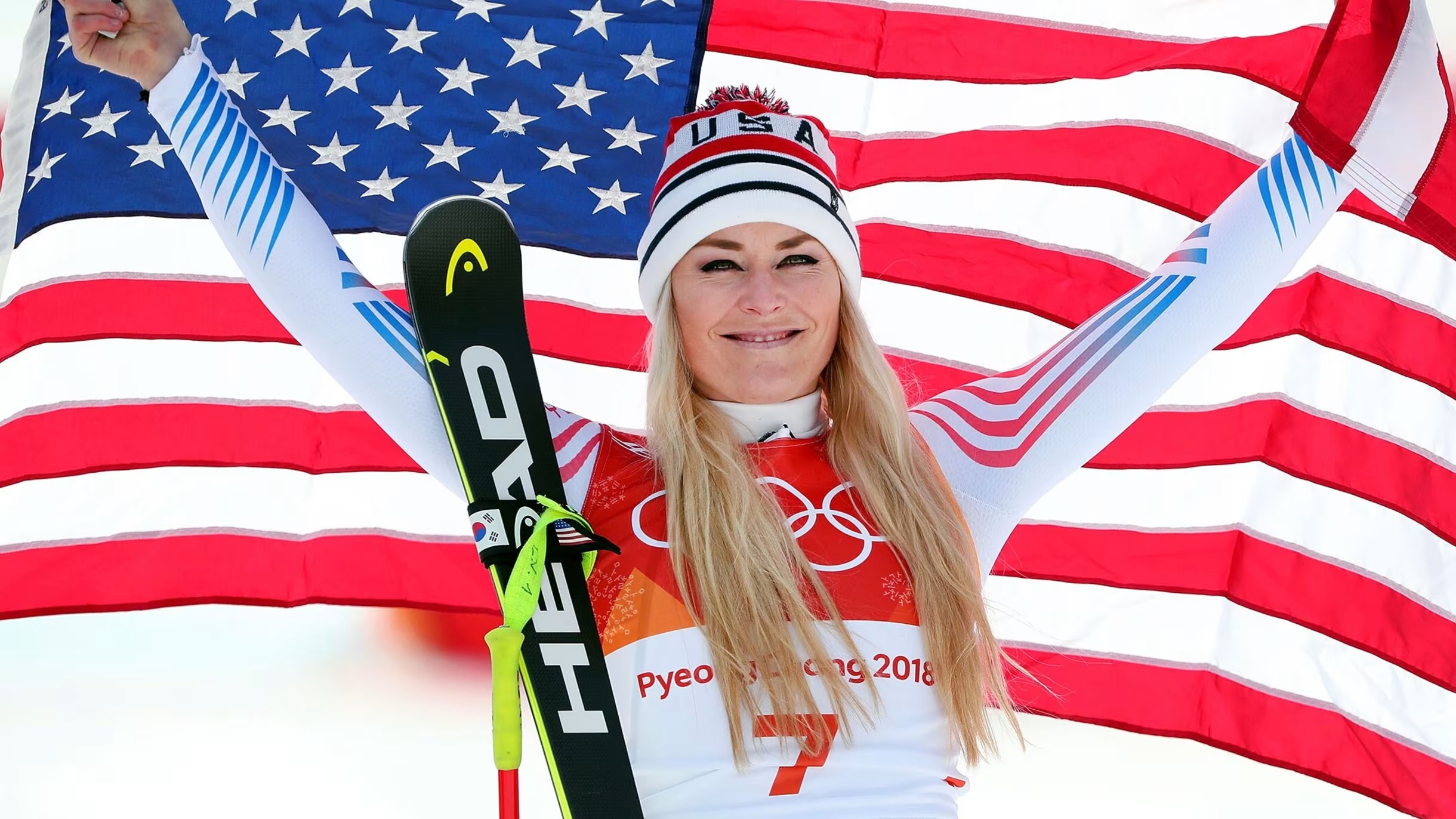 Lindsey Vonn, an American skier with long blonde hair, wears a Team USA beanie and uniform while holding up an American flag with her skis resting against her arm.