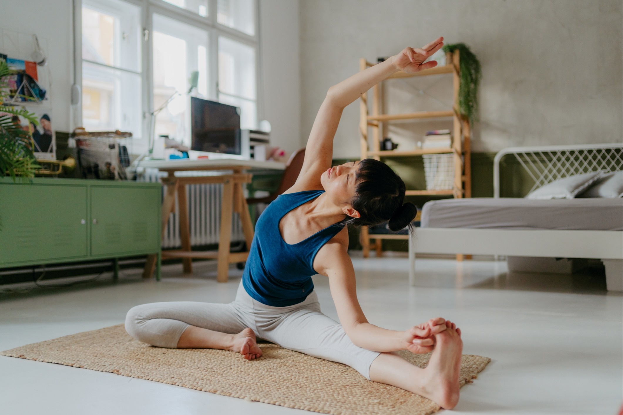 Person stretching on a mat in a bright bedroom, leaning into a side stretch with one arm raised and the other holding their foot, sunlight coming through a window behind them.