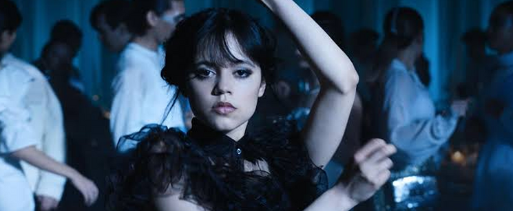 Jenna Ortega performing her iconic dance scene as Wednesday Addams in a dark room surrounded by school party guests.