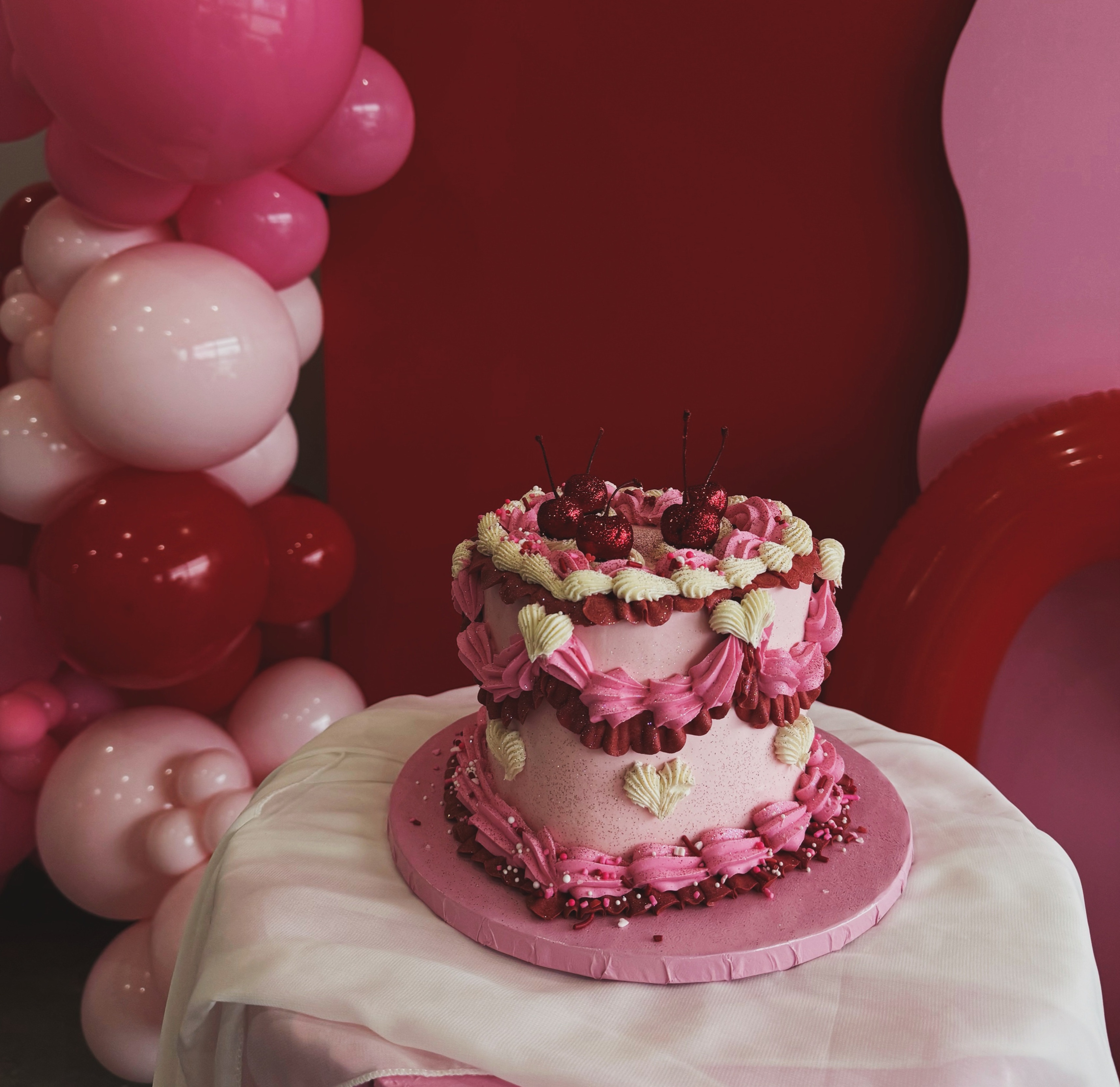 Pink frosted cake with cherries on top displayed on a table, surrounded by pink and red balloon decorations.