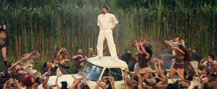 Bad Bunny in an all-white outfit standing on top of a vintage car surrounded by a crowd during a live performance.