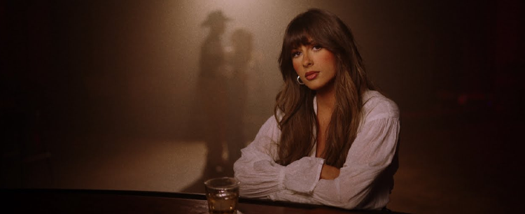 Singer Ella Langley sitting at a dimly lit bar with a glass of whiskey, leaning her arms on the counter.