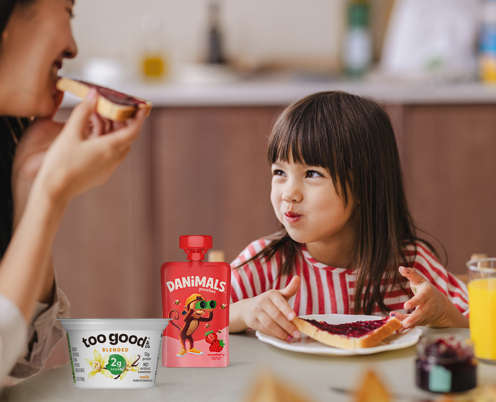 Girl smiling at adult while eating toast at a table in the kitchen with Danimals and too good products.