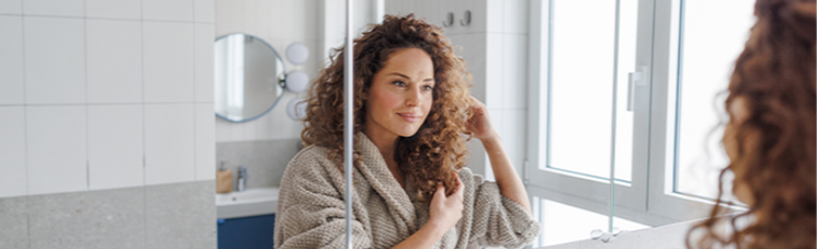 Calm moment of a midlife woman in a modern bathroom, wearing a robe and looking into the mirror. A scene of self-care, skincare, and everyday wellness.