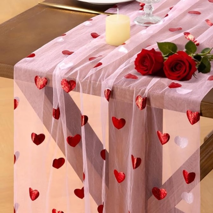 A pink tulle table runner with red hearts on it sits on a table with 2 red roses and a candle.