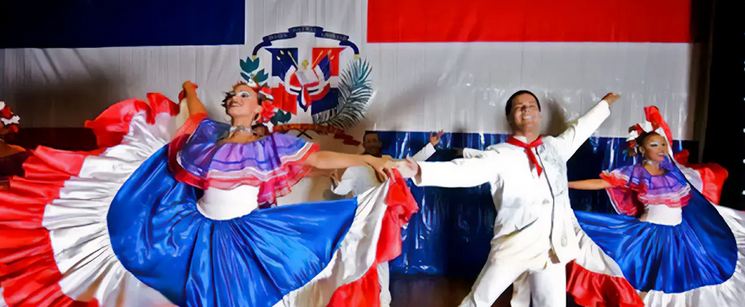 Dancers performing in front of the Dominican Republic flag in vibrant blue and red dresses alongside a male dancer in white.