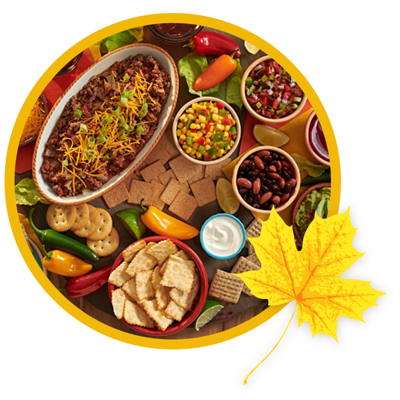 An overhead spread of tailgate snacks including a dish of chili with shredded cheese, crackers, salsa, black beans, guacamole, sour cream, corn salsa, and fresh peppers, next to a fall leaf graphic.