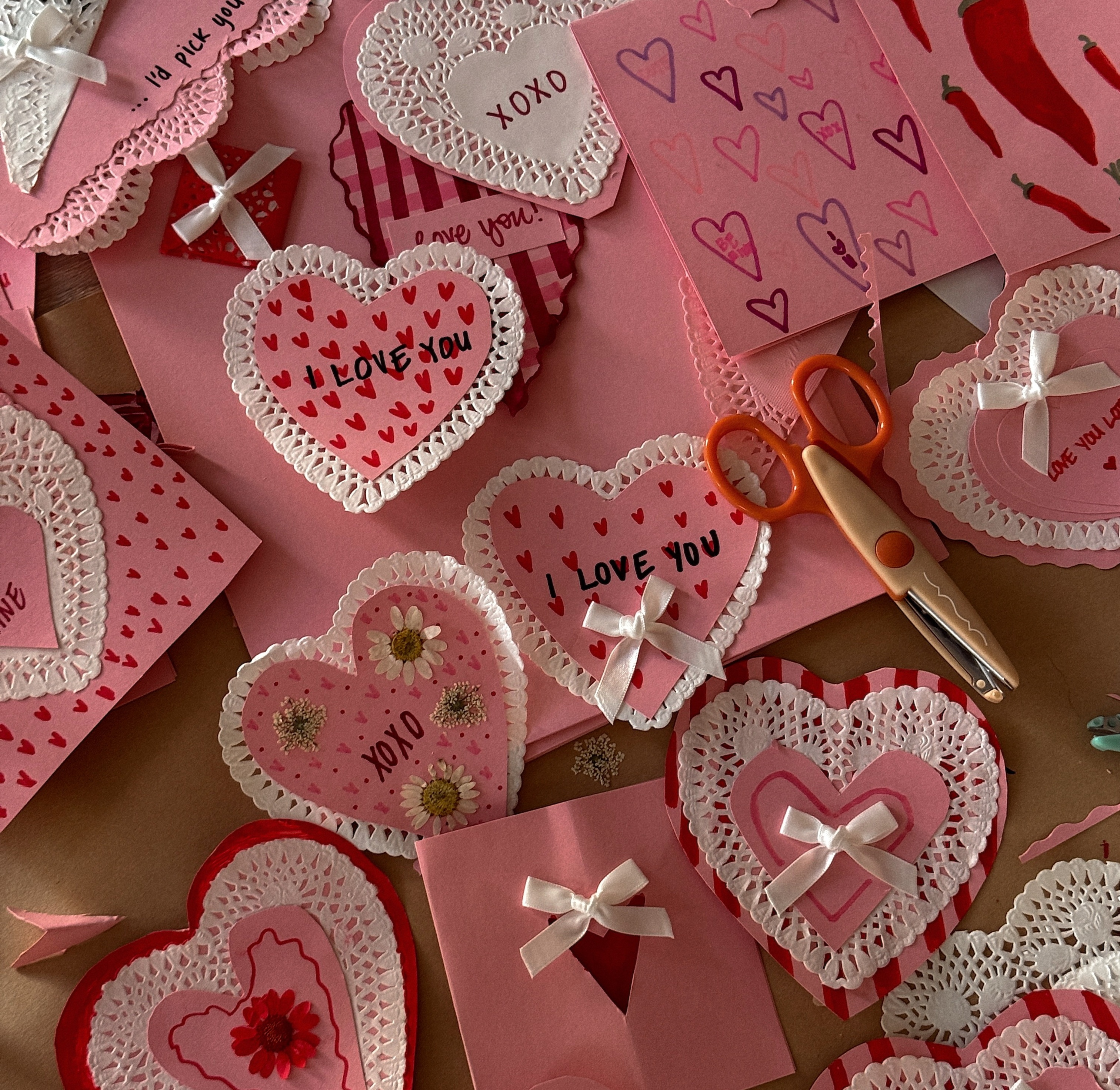 Pink and red handmade Valentine’s cards with heart shapes, paper lace details, ribbons, and scissors spread across a table.