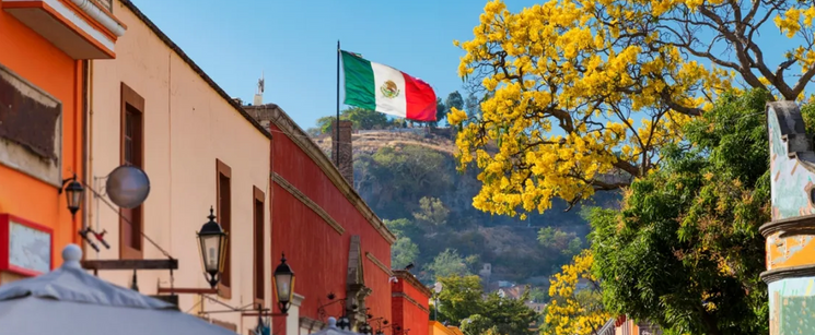 A scenic street view in Mexico with a Mexican flag flying over traditional colorful buildings and yellow blossom trees.