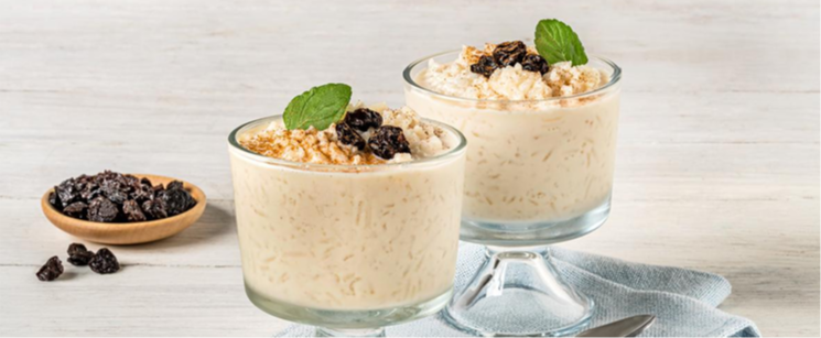 Two creamy rice-based desserts served in glass dishes, topped with cinnamon, raisins, and fresh mint, with a bowl of raisins beside them. / Dos postres cremosos a base de arroz servidos en recipientes de vidrio, cubiertos con canela, pasas y menta fresca, con un tazón de pasas al lado.