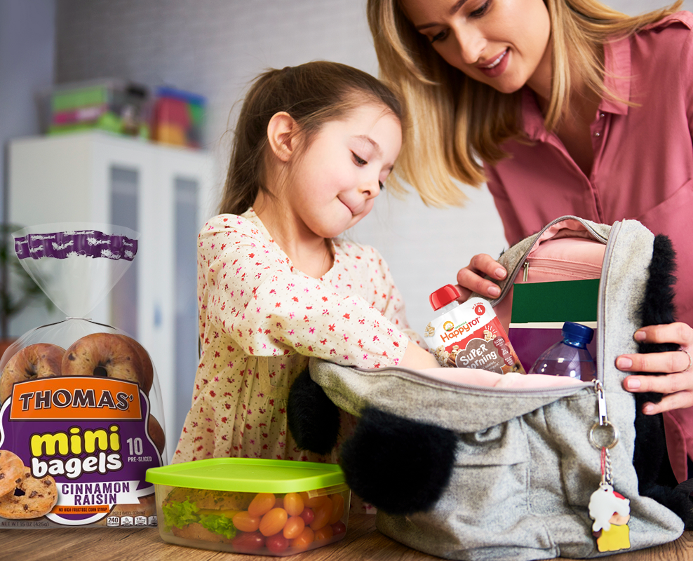 Girl and parent packing a school bookbag with snacks and bagels on the table.