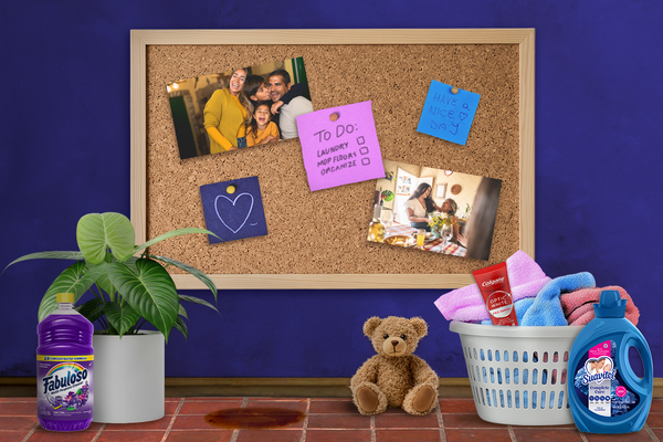 A board with family photos and to-do lists on a blue wall with Spanish tiles. On the floor to the left, there is a plant and Fabuloso. On the right, there is a basket of unfolded laundry, Colgate, and Suavitel. In the middle, there is a spill on the floor and a teddy bear. / Un tablero con fotos de familia y listas de tareas en una pared azul con azulejos españoles. En el piso a la izquierda hay una planta y Fabuloso. A la derecha está una canasta de ropa sin doblar, Colgate y Suavitel. En el medio hay un derrame en el piso y un osito de peluche.