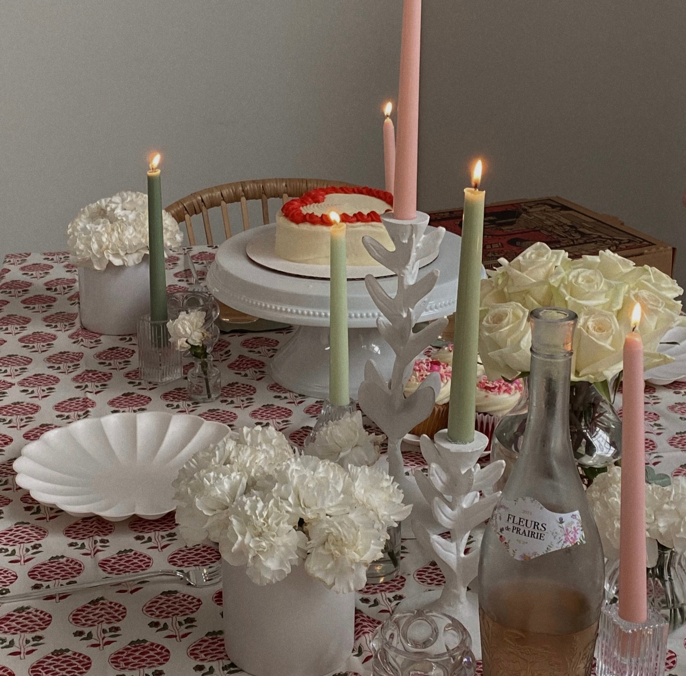 Decorated table with lit candles, white flowers, cakes on stands, and pastel tableware arranged for a cozy gathering.