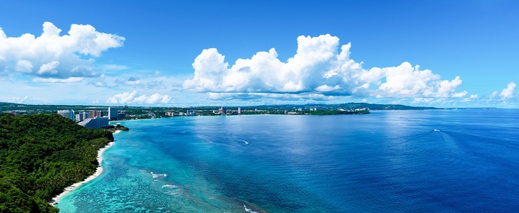 An aerial panorama of a tropical coastline with vibrant turquoise water, a sandy beach, and lush green hills under a bright blue sky.