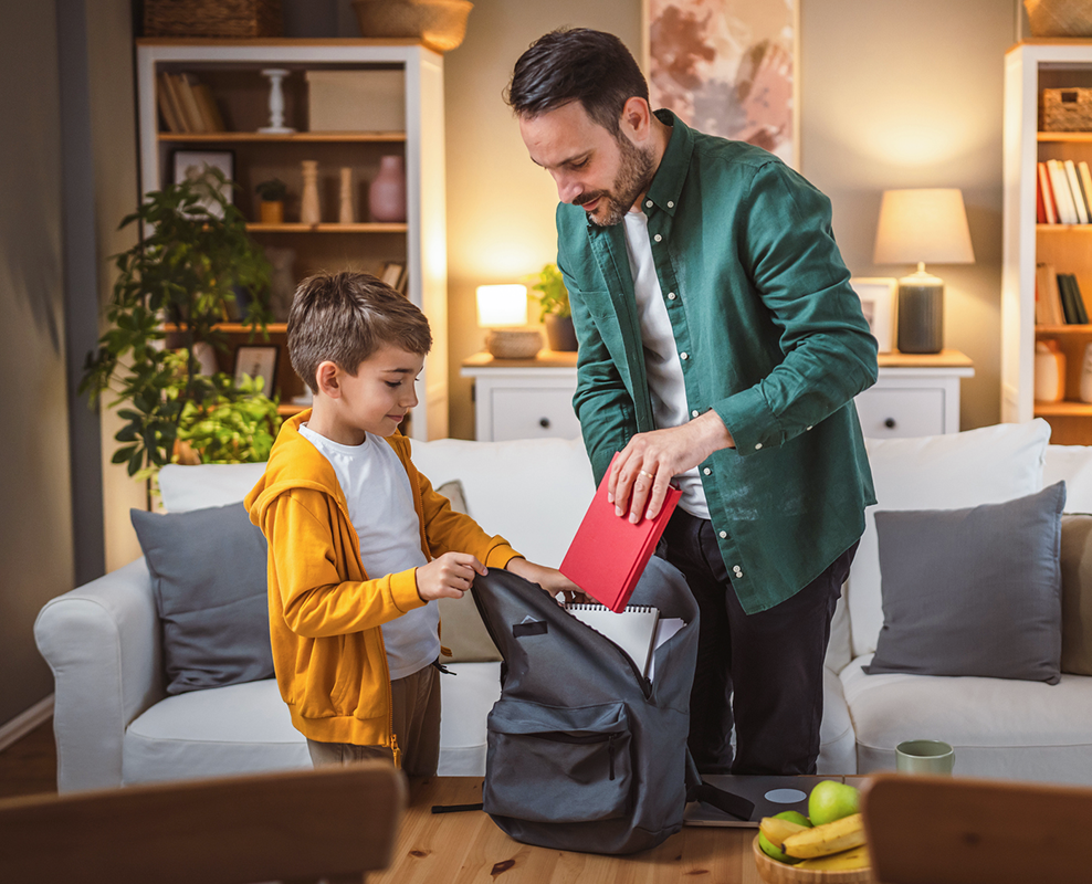 Boy and parent packing a school bookbag on a coffee table in the living room.