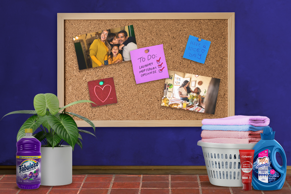 A board with family photos and to-do lists on a blue wall with Spanish tiles. On the floor to the left, there is a plant and Fabuloso. On the right, there is a laundry basket, Colgate, and Suavitel. / Un tablero con fotos de familia y listas de tareas en una pared azul con azulejos españoles. En el piso a la izquierda hay una planta y Fabuloso. A la derecha está una canasta de ropa, Colgate y Suavitel.